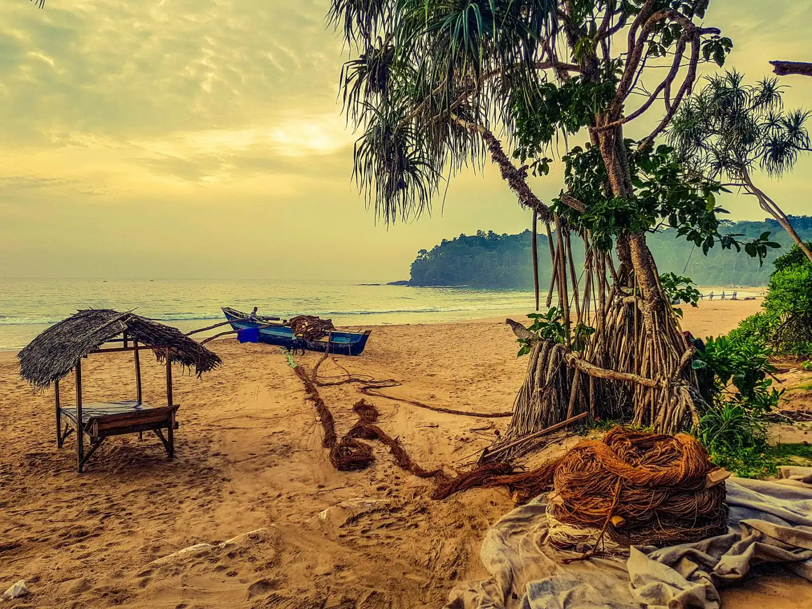 Talalla beach with a thatched shelter, a blue fishing boat, coiled ropes and a screw pine tree under a hazy golden sky.