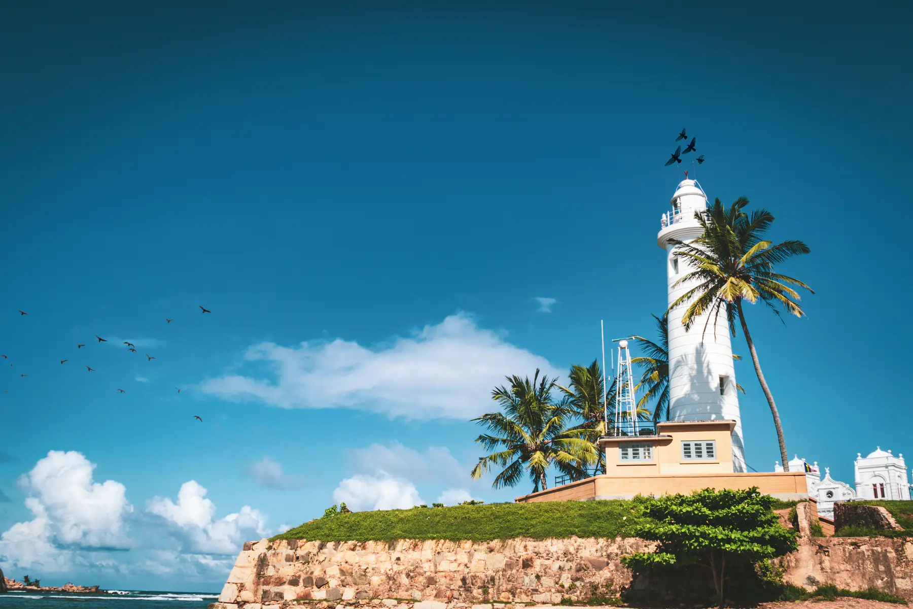 Galle Lighthouse on the fort wall under a blue sky, with palms and birds, on the south-west coast that is in season in December.