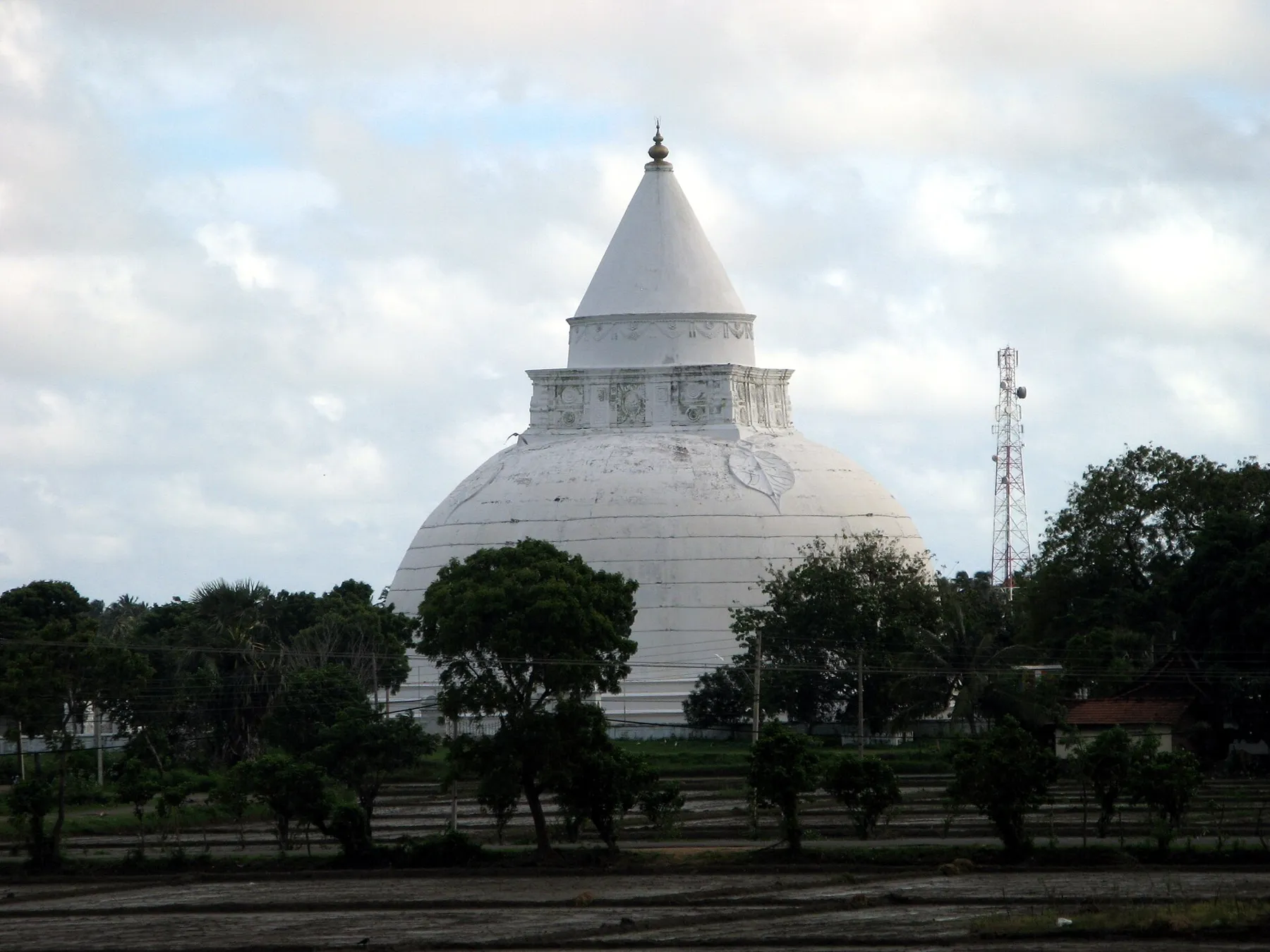 The large white dome and conical spire of the Tissamaharama dagoba under a cloudy sky, with trees and paddy fields in front.