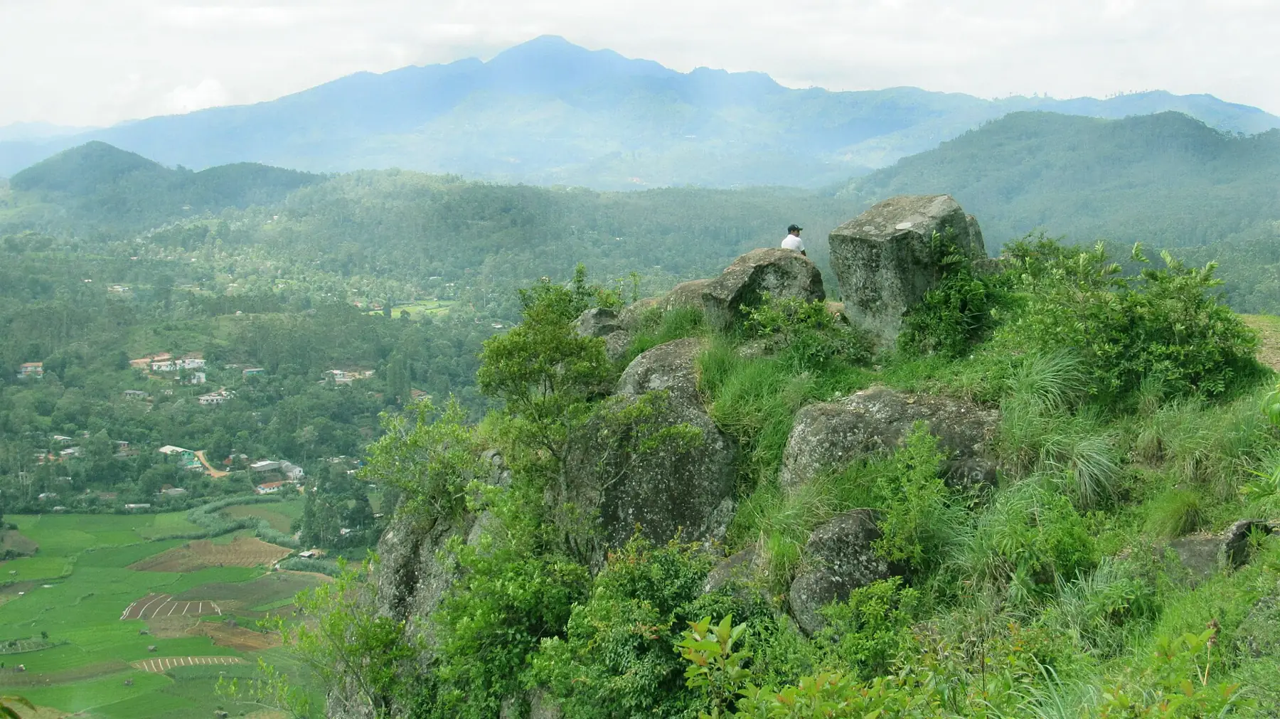 A person sits on a rocky outcrop at Porawagala above a green valley of villages and farmland, with misty mountains beyond.