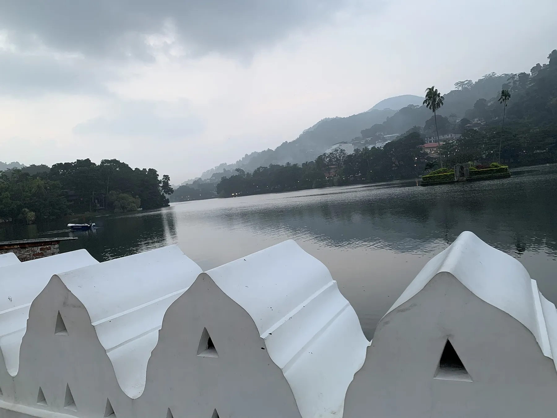 Kandy Lake behind its white scalloped wall, with palm trees on a small island and wooded hills beyond.