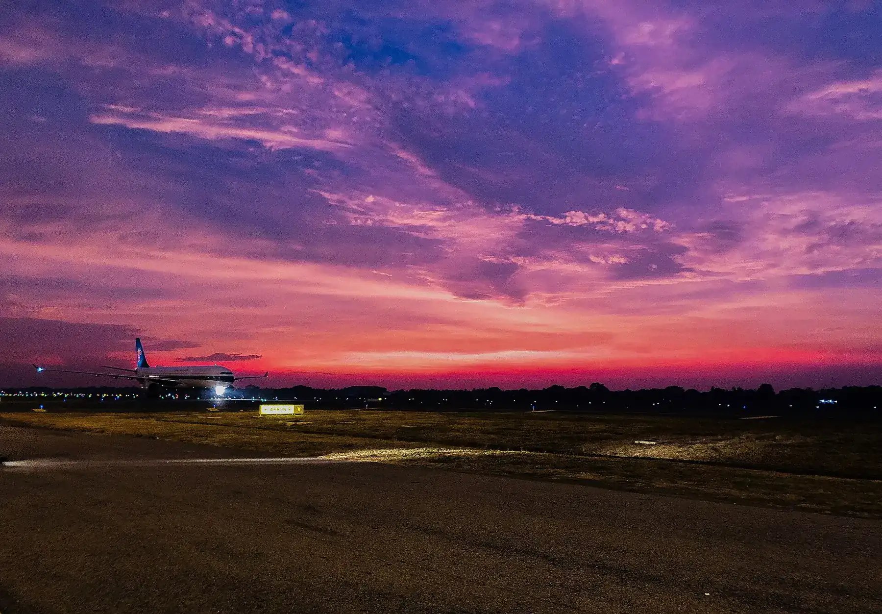 A purple and orange sunset sky over the runway at Bandaranaike International Airport, with an aircraft in the distance.