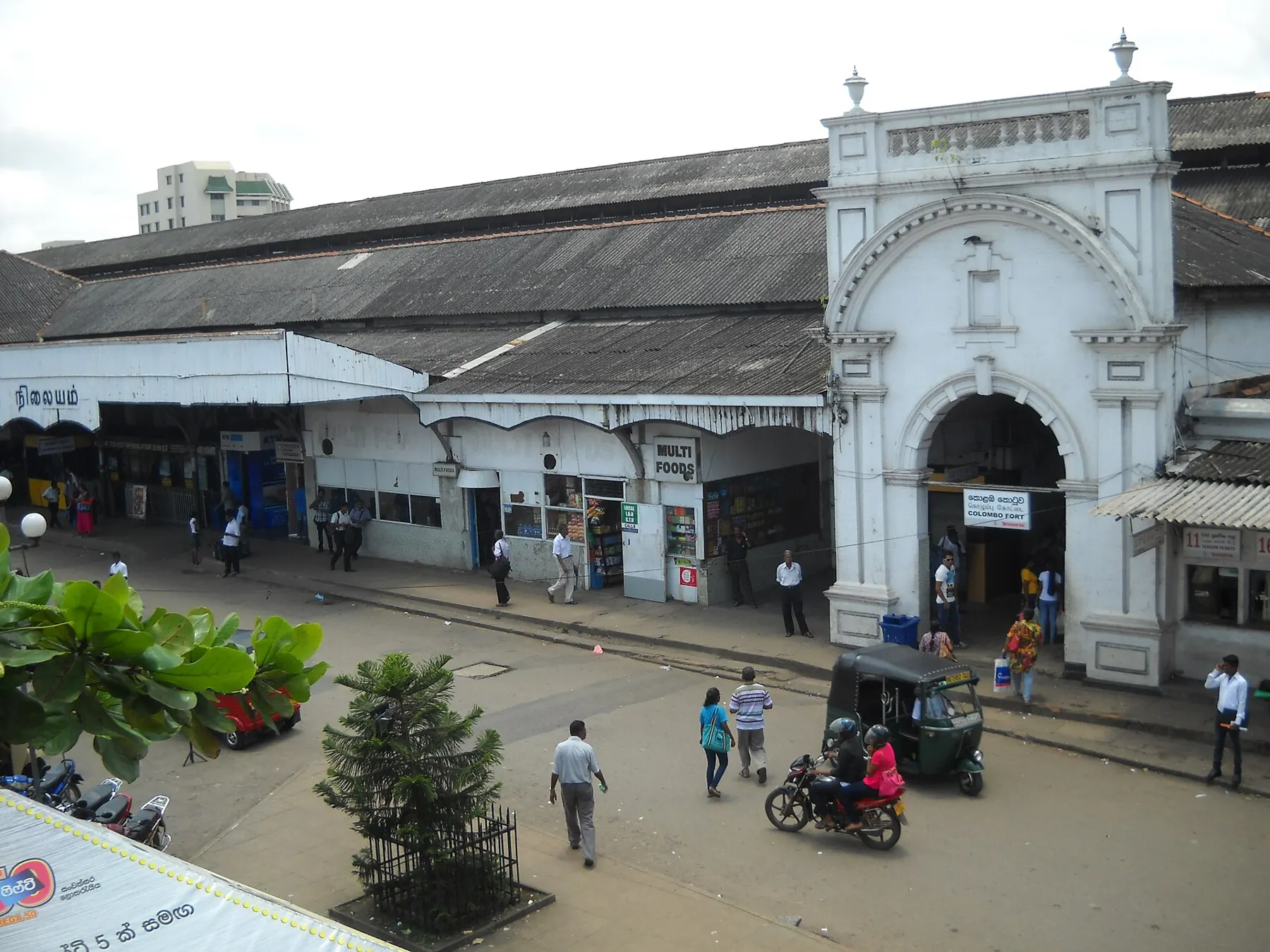 The white arched entrance of Colombo Fort railway station, with travelers, a tuk tuk and a motorbike out front.