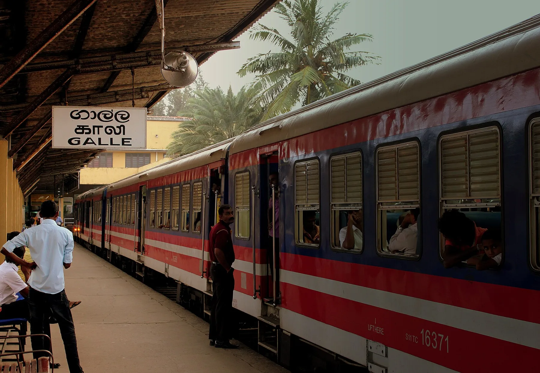 A red and blue Sri Lanka Railways train at Galle station, with the platform sign and palm trees beyond.