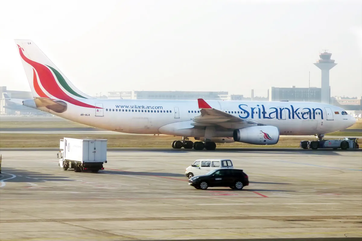 A SriLankan Airlines Airbus A330 on an airport apron, with a service truck and cars in front and a control tower behind.